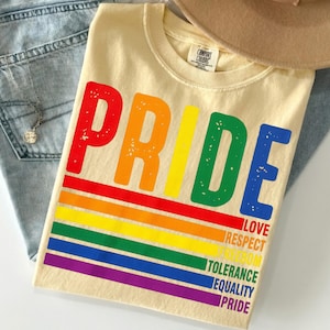Könnte beinhalten: Cremefarbenes T-Shirt mit dem Wort "PRIDE" in Regenbogenfarben. Unter dem Wort "PRIDE" befindet sich ein Regenbogenstreifen-Design. Das Shirt zeigt auch die Wörter "LOVE, RESPECT, FREEDOM, TOLERANCE, EQUALITY, PRIDE".