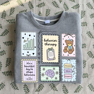May include: A heather gray crewneck sweatshirt featuring a graphic print of six rectangular frames. The frames contain illustrations of a bar chart, a clipboard with the text behavior therapy, a teddy bear, the text it's a beautiful day to shape behaviors, a brain, and a flow chart labeled Antecedent, Behavior, and Consequence.