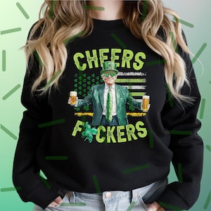 May include: Black sweatshirt with a graphic featuring a man in a green suit, hat, and sunglasses holding two mugs of beer. The text "CHEERS F*CKERS" is displayed in green, with a shamrock and an American flag design.