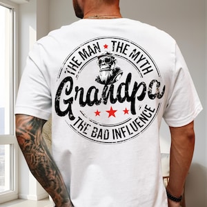 以下が含まれることがあります： 背中に円形のグラフィックが入った白いTシャツ。グラフィックには、サングラスとひげを生やした漫画のゴリラとともに、黒色の「THE MAN THE MYTH Grandpa THE BAD INFLUENCE」というテキストが表示されています。