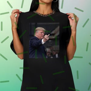 May include: Black t-shirt featuring a photographic print of a man in a suit gesturing with his middle finger. The image is centered on the shirt. The person wearing the shirt is also wearing a gold chain necklace.