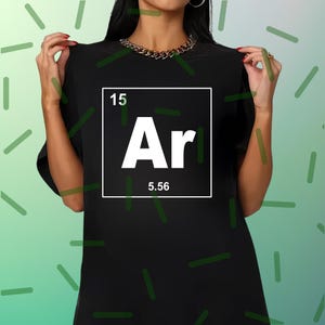 Op de afbeelding: Zwart T-shirt met een witte vierkant met het elementsymbool "Ar" en het getal "5,56" eronder. Het getal "15" staat boven het symbool. Het shirt heeft een eenvoudige ronde hals.