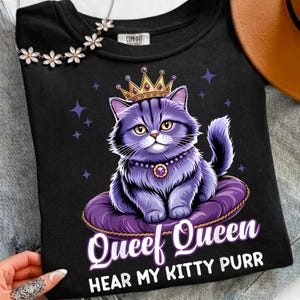 May include: Black crew neck t-shirt featuring a purple cat wearing a gold crown and a pearl necklace. The cat is sitting on a purple cushion. The text "Queef Queen Hear My Kitty Purr" is printed below the cat.