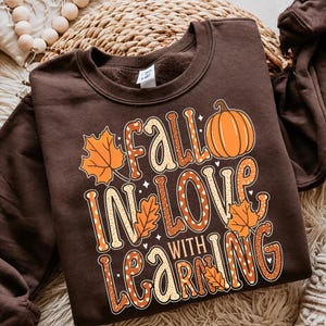 May include: A dark brown crewneck sweatshirt featuring a graphic print with the text "FALL IN LOVE WITH LEARNING" in orange and white patterned lettering, accompanied by illustrations of a bright orange pumpkin and several orange maple leaves.