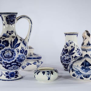 Könnte beinhalten: Eine Sammlung von weiß-blauer Delfter Keramik. Die Stücke umfassen einen Krug, zwei Vasen, eine Schale mit Deckel und eine kleine Schale. Die Designs zeigen Blumenmuster und eine Szene mit Segelboot und Windmühle.