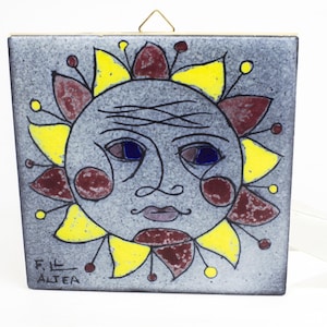 May include: Square ceramic tile featuring a sun face design. The sun has a grey face with blue eyes, a moustache, and red cheeks. Yellow and maroon rays surround the face. The tile has a brass hanger and the text "F.L. ALTEA".