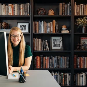 May include: A woman with blonde hair and glasses smiles in front of a computer. Behind her is a dark bookshelf filled with books, framed pictures, and decorative items. The scene suggests a home office or study.