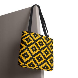 May include: A tote bag with a geometric pattern in black and yellow. The bag has black handles and is hanging against a gray surface. The pattern is a repeating diamond design.