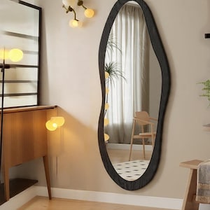 May include: A large, organically shaped mirror with a dark, textured frame hangs on a beige wall. The mirror reflects a room with a wooden chair, a rug, and a window with white curtains. The room is lit by warm-toned lights.