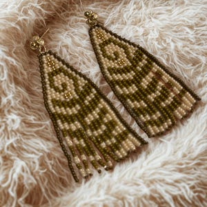 May include: A pair of beaded earrings in olive green and cream, featuring a geometric design. The earrings have a fringe and gold-coloured ear wires. They are presented on a fluffy, cream-coloured surface.