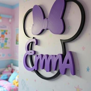 May include: A wall-mounted sign featuring a black circular outline shaped like mouse ears with a purple bow at the top and the name Emma in purple lettering across the centre. The text MINNIE VERSION 2 is printed above the sign.