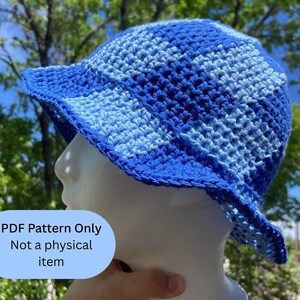 May include: A blue and light blue crochet bucket hat. The hat is displayed on a mannequin head. The text "PDF Pattern Only Not a physical item" is in a light blue box in the lower left corner. The hat has a brim and is made of a textured yarn.