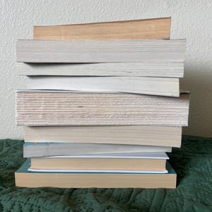 May include: A stack of eight books, mostly white with a few brown covers, on a green patterned surface.