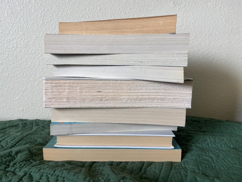 May include: A stack of eight books, mostly white with a few brown covers, on a green patterned surface.
