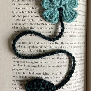 May include: A close-up of a green and blue crocheted flower bookmark. The bookmark is lying on a book with text about a character named Ithan.