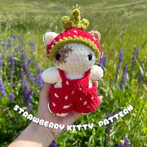 May include: A handmade crochet strawberry kitty plushie. The plushie is white with brown spots, wearing a red strawberry hat and overalls with white polka dots. The text "STRAWBERRY KITTY PATTERN" is displayed on the image.