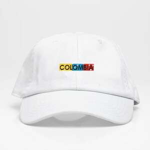 May include: White baseball cap with a curved brim. The word "COLOMBIA" is displayed across the front in a colorful, rectangular design. The letters are yellow, blue, and red, representing the colors of the Colombian flag.