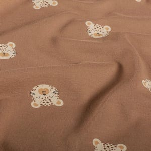 Könnte beinhalten: Nahaufnahme eines braunen Stoffes mit einem sich wiederholenden Muster aus lächelnden Leoparden-Gesichtern. Die Leoparden-Gesichter sind weiß mit braunen Flecken, beigen Ohren und einer beigen Nase. Der Stoff hat ein weiches, strukturiertes Aussehen.