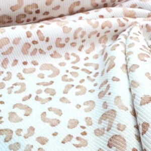 Könnte beinhalten: Nahaufnahme eines weißen Stoffes mit einem sich wiederholenden Muster aus braunen Leopardenflecken. Der Stoff ist gefaltet und zeigt Textur und Tiefe. Das Leopardenmuster ist ein klassisches und vielseitiges Muster.