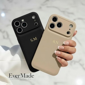 Puede incluir: Dos fundas para smartphone, una negra y otra beige, con acabado texturizado y recorte elevado para la cámara. La funda negra tiene grabadas las iniciales doradas S.M, mientras que la beige muestra las iniciales doradas A.N. El texto EverMade es visible.