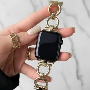 May include: A black smartwatch with a gold-toned metal link band featuring circular and rectangular connectors. The watch is accompanied by two gold-toned rings, one with a cage-like design and the other a simple polished band.