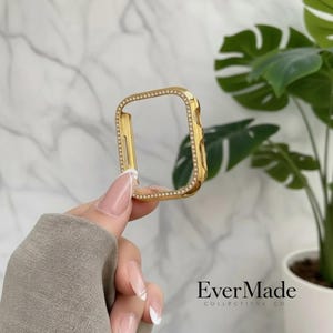 May include: A gold-toned rectangular watch case cover featuring a border of small, clear embedded crystals. The frame has precise cutouts along the sides for buttons and ports, designed to fit over the body of a digital smartwatch device.