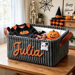 May include: A rectangular dark gray corduroy storage basket with brown leather handles. The name Julia is embroidered in orange yarn on the front, alongside a white ghost patch. Inside are a black and orange pumpkin onesie, candy, and a pumpkin.