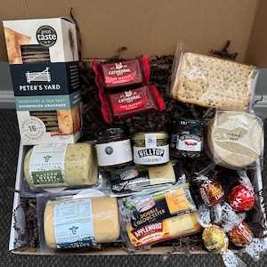 May include: A food hamper containing a selection of gourmet treats. The contents include crackers, cheese, honey, jam, and chocolates. Brands such as Peter's Yard, Cathedral City, and Ilchester are visible. The items are presented in a cardboard box.