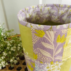 May include: A cylindrical fabric storage basket with a patchwork design. The basket is made of different fabrics with floral patterns in shades of green, purple, and white.