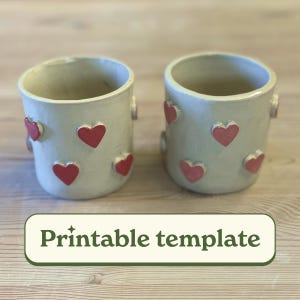 May include: Two white ceramic mugs with red heart designs. The mugs are on a wooden surface. Printable template.
