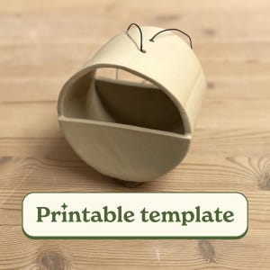 May include: A beige ceramic hanging planter with a rounded shape and a small opening at the top for hanging. The planter has a curved, half-circle opening on the front for planting.