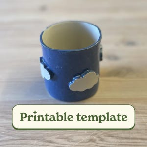 Puede incluir: Una taza de cerámica azul con detalles en forma de nube blanca. La taza está sobre una superficie de madera. Plantilla imprimible.
