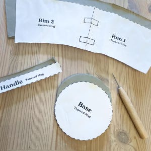 May include: A craft project with paper templates for a tapered mug. The templates include "Rim 2", "Rim 1", "Handle", and "Base". A wooden-handled tool and a gray clay circle are also present on a wooden surface.