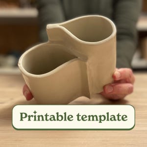 Könnte beinhalten: Ein weißer Keramik-Besteckhalter mit zwei Fächern. Der Halter ist in Form von zwei miteinander verbundenen Tassen geformt. Der Text "Druckbare Vorlage" ist unter dem Halter sichtbar.