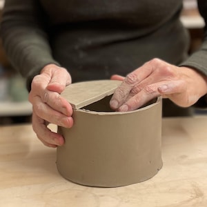 May include: A person is making a ceramic lid for a cylindrical container. The lid is a light brown color and is being placed on top of the container. The container is also light brown and has a smooth surface.