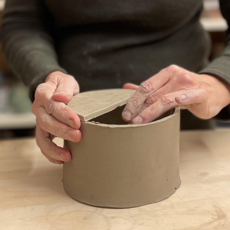 May include: A person is making a ceramic lid for a cylindrical container. The lid is a light brown color and is being placed on top of the container. The container is also light brown and has a smooth surface.