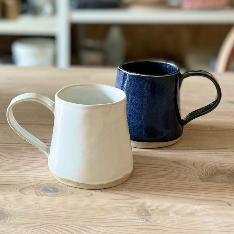 May include: Two ceramic mugs on a wooden surface. One is white with a beige base and handle, the other is a dark blue with a black handle. The mugs are of different sizes and shapes, suggesting a handmade or artisanal quality.