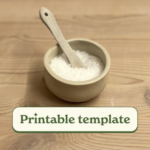 Puede incluir: Un salero de cerámica blanca con una pequeña cuchara de madera en su interior. El salero está lleno de cristales de sal blancos.