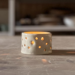 May include: White ceramic candle holder with a textured surface and small holes throughout. The candle holder is lit and casting a warm glow.