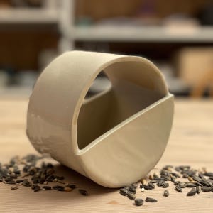 May include: A white ceramic bird feeder with a rounded shape and a cut-out opening. The feeder is sitting on a wooden surface with scattered birdseed.