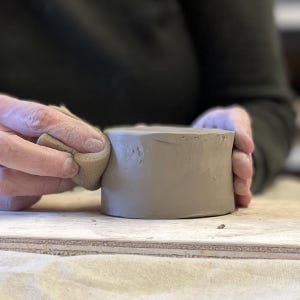 May include: A person's hands are smoothing the surface of a gray clay pot with a damp sponge.
