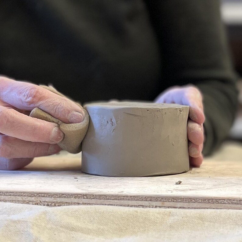 May include: A person's hands are smoothing the surface of a gray clay pot with a damp sponge.