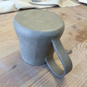 May include: A handmade, unglazed ceramic mug with a rounded body and a large, curved handle. The mug is a light gray color and sits on a wooden surface. The mug is likely for drinking beverages.