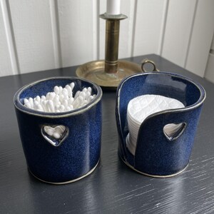 May include: Two matching navy blue ceramic containers with heart-shaped cutouts. One holds cotton swabs, the other holds cotton pads. The containers are on a dark surface, with a brass candlestick in the background.