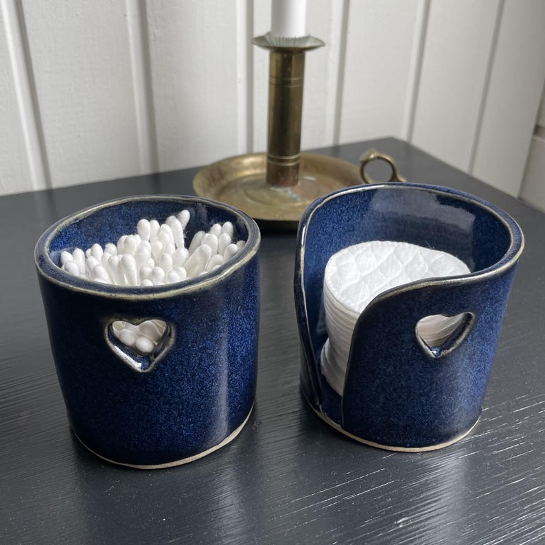 May include: Two matching navy blue ceramic containers with heart-shaped cutouts. One holds cotton swabs, the other holds cotton pads. The containers are on a dark surface, with a brass candlestick in the background.