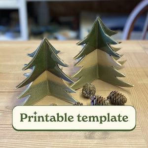 May include: Two ceramic Christmas tree decorations with a green and gold glaze, displayed on a wooden table with pine cones. The trees have a layered design. The image includes a banner with the text "Printable template".