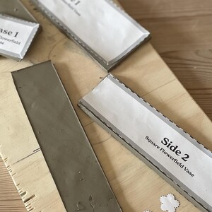 May include: A close-up of a gray clay tile with a white label that reads "Side 2 Square Flowerfield Vase".
