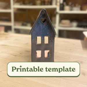以下が含まれることがあります： ハート型の切り抜きと4つの長方形の窓が付いた、家型のセラミックキャンドルホルダー。家は光沢のある仕上げの濃い青色です。「Printable template」というテキストが緑色のバナーに表示されています。