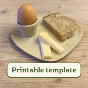 Peut inclure: Une assiette de petit-déjeuner avec un œuf à la coque dans un coquetier en céramique, deux bâtonnets de fromage et une tranche de pain. L'assiette est en forme de cœur et porte le texte "Printable template".