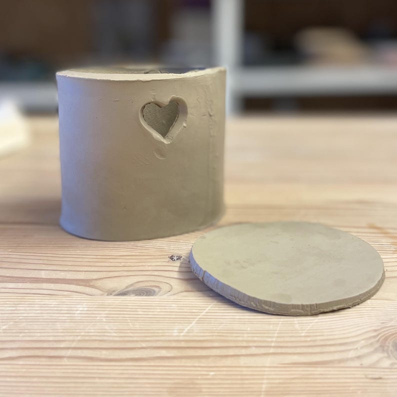 May include: A handmade, cylindrical clay container with a heart-shaped cutout. The container is light gray and has a matching, round lid. The items are displayed on a wooden surface. The container is approximately 4 inches tall.